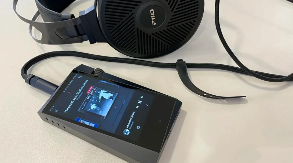 Headphones connected to a digital audio player displaying an album cover and track information on a light-colored surface. For soundhub's blog Best  Audio Accessories.