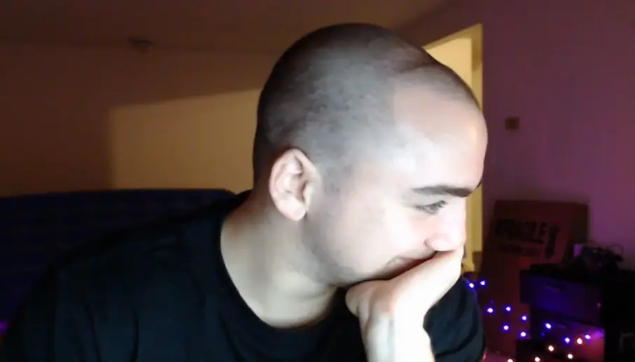 Person with a shaved head, resting chin on hand, looks to the side. Background features dim lighting and string lights.