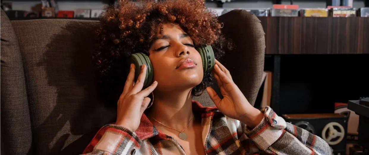 Person with curly hair wearing plaid shirt, enjoying music with large green headphones, eyes closed and relaxed in a cozy room.